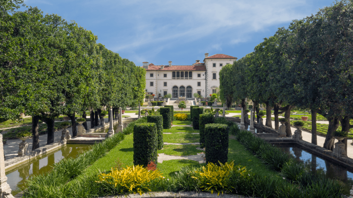 Vizcaya’s exterior presents an 18th-century Italian villa façade in the Veneto style, with white stucco covering the limestone construction, under a terracotta barrel-shaped tile roof. Corner towers feature low-pitched hipped roofs covered in the same barrel-shaped tiles. Three arched center-point windows and a columned second-floor balcony overlook the formal gardens. The gardens, designed by landscape architect Diego Suarez (1888–1974), are characterized by flanking rows of manicured oaks, geometric-shaped hedges, reflecting pools, statuary, and topiary. (Robin Hill/Vizcaya Museum and Gardens)