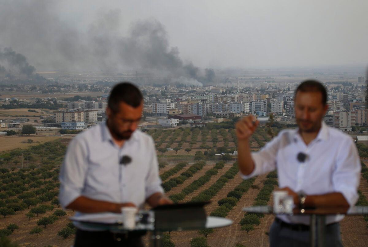 TV journalists talk during a live broadcast on a hilltop in Ceylanpinar, Sanliurfa province, southeastern Turkey, as in the background smoke billows from targets in Ras al-Ayn, Syria, during bombardment by Turkish forces on Oct. 16, 2019. (AP Photo/Lefteris Pitarakis)