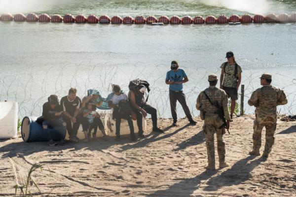 Asylum-seeking illegal immigrants sit by concertina wire fencing while waiting to be transported by U.S. law enforcement officers after crossing the Rio Grande into the U.S. from Mexico in Eagle Pass, Texas, on July 24, 2023. (Go Nakamura/Reuters)