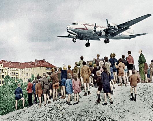 At Tempelhof Airport, Berliners watching a C-54 land. The aircraft was used in the Berlin Airlift of 1948–49, which defied the Soviet blockade by delivering supplies to the city's western sectors. (Public Domain)